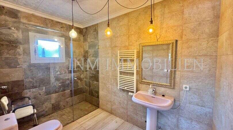 Badezimmer der Finca in Llucmajor mit Natursteinoptik, Glasdusche, Hängelampen und goldenem Spiegel.