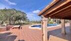 Überdachte Terrasse mit Pool und weitläufigem Garten der Finca in Llucmajor unter blauem Himmel.