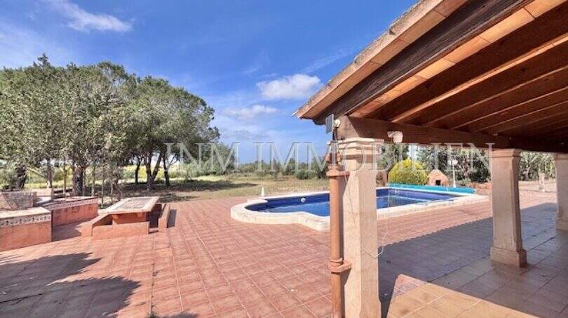 Überdachte Terrasse mit Pool und weitläufigem Garten der Finca in Llucmajor unter blauem Himmel.