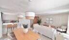 Luminosa sala de estar-comedor con moderna mesa de madera, sofás blancos y cocina abierta en la casa de nueva construcción en Puig de Ros