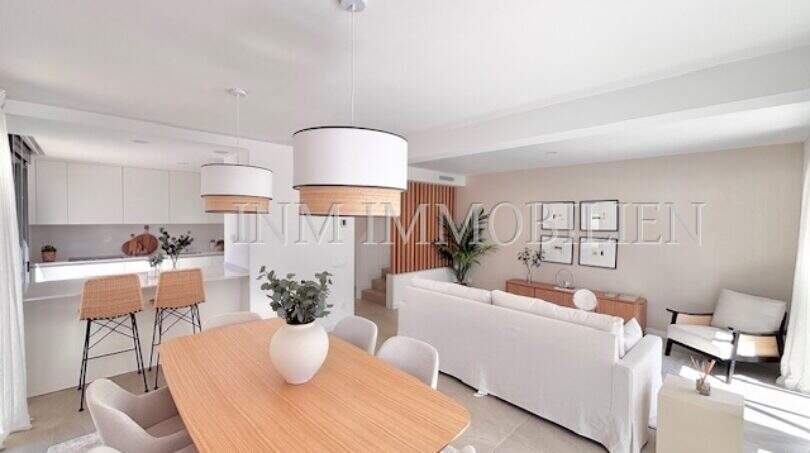Luminosa sala de estar-comedor con moderna mesa de madera, sofás blancos y cocina abierta en la casa de nueva construcción en Puig de Ros