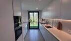 Cocina moderna empotrada con frentes blancos, electrodomésticos de vitrocerámica y vista al jardín en la casa de nueva construcción Puig de Ros