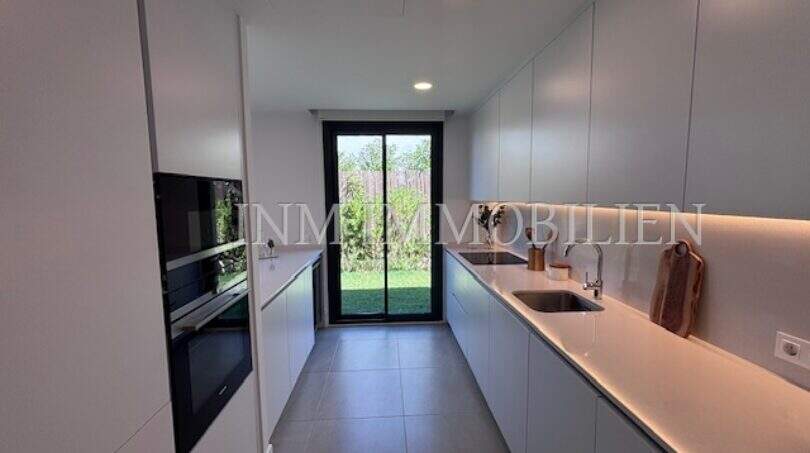 Cocina moderna empotrada con frentes blancos, electrodomésticos de vitrocerámica y vista al jardín en la casa de nueva construcción Puig de Ros