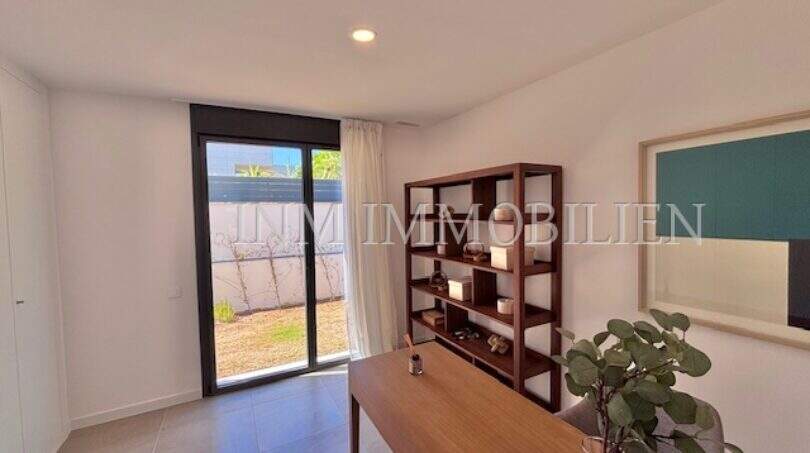 Moderno despacho con escritorio de madera, pared de estanterías y puerta corredera de cristal al soleado jardín privado en la casa de nueva construcción Puig de Ros.