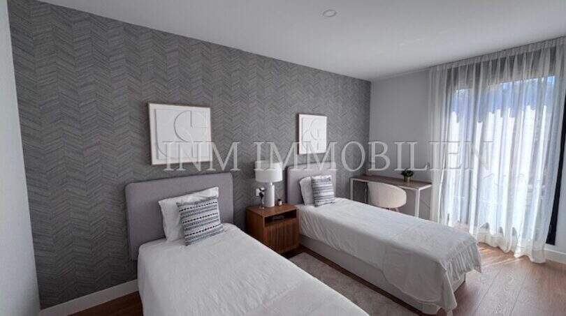 Dormitorio luminoso con dos camas individuales, papel pintado gris estampado y suelo de parqué en la casa de nueva construcción Puig de Ros