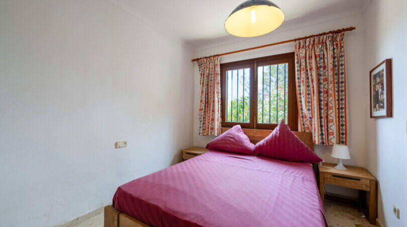 Dormitorio de la villa de Cala Murada con cama de matrimonio, muebles de madera y cortinas de colores en la ventana enrejada.