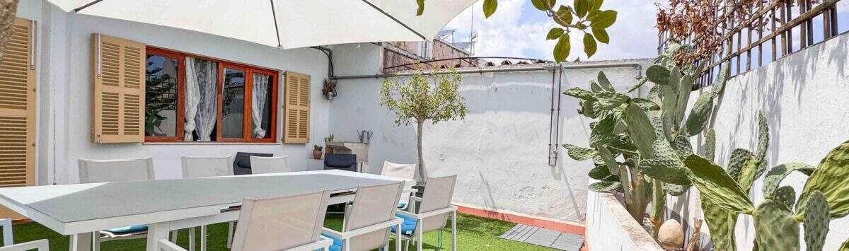 Terraza soleada con muebles de jardín blancos, sombrilla, zona de césped artificial y cactus en Felanitx