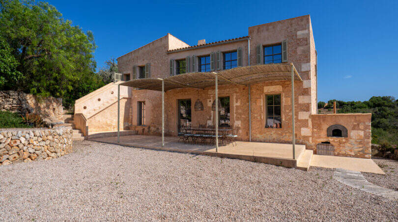Außenansicht einer rustikalen Sandstein-Finca mit überdachter Terrasse, Außenkamin und Kiesvorplatz in Cales de Mallorca.