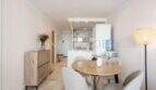 Comedor luminoso con mesa de madera redonda, sillas modernas y cocina abierta en apartamento en Port Adriano, Calvià.