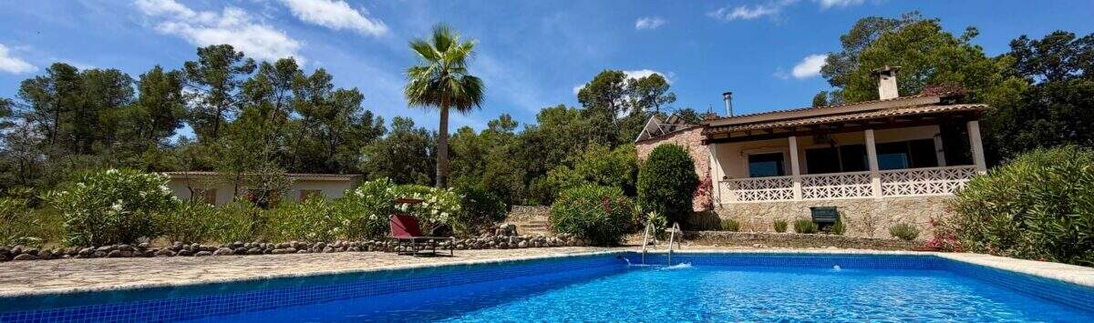 Großer blauer Pool vor rustikaler Finca mit Terrasse, Palme und Pinienwald unter blauem Himmel in Sant Joan