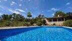 Großer blauer Pool vor rustikaler Finca mit Terrasse, Palme und Pinienwald unter blauem Himmel in Sant Joan