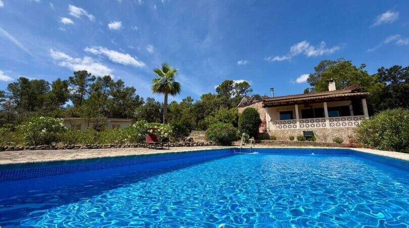Großer blauer Pool vor rustikaler Finca mit Terrasse, Palme und Pinienwald unter blauem Himmel in Sant Joan