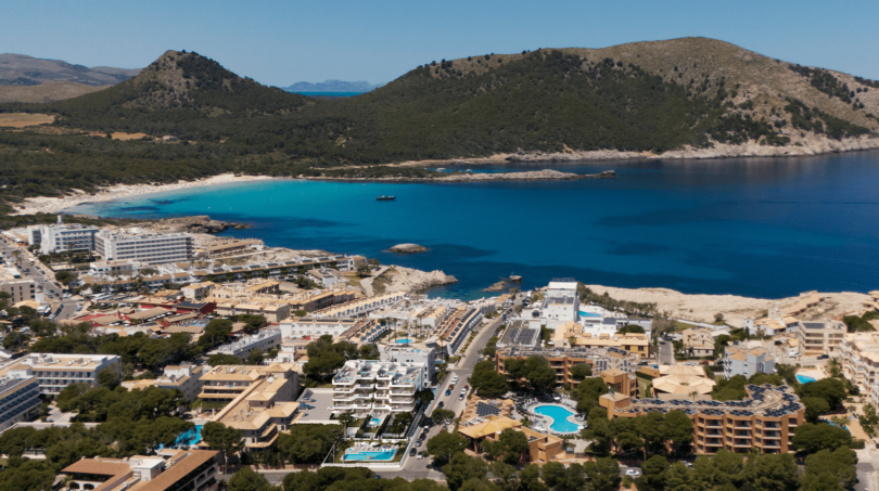Luftaufnahme von Cala Ratjada mit türkisblauem Meer, Sandstrand und Hotelanlage, umgeben von grünen Hügeln.