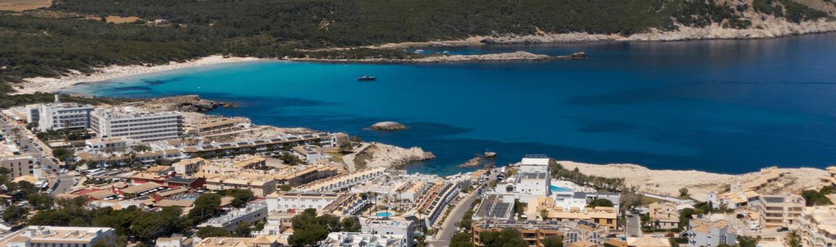 Luftaufnahme von Cala Ratjada mit türkisblauem Meer, Sandstrand und Ferienanlage, umgeben von grünen Hügeln.