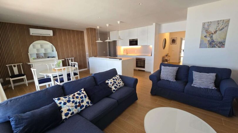 Helles Wohnzimmer mit blauen Sofas, offener Küche und Holzwandverkleidung im Hafenapartment Portocolom