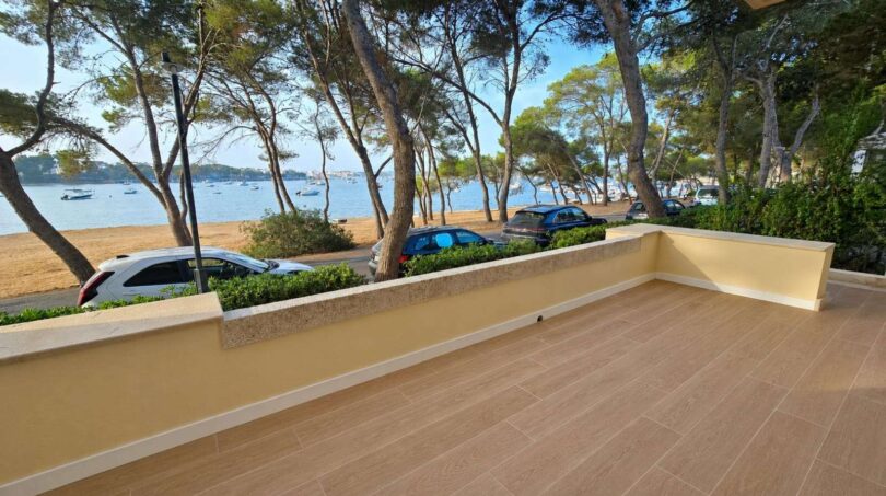 Sonnige Terrasse mit Holzfliesenoptik, Meerblick auf Hafenbucht von Portocolom, Pinien und Sandstrand direkt davor.