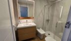 Modernes Badezimmer mit Marmorwänden, Holzwaschtisch, beleuchtetem Spiegel und Glasdusche im Hafenapartment Portocolom