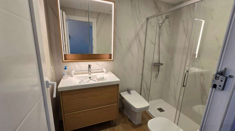 Modernes Badezimmer mit Marmorwänden, Holzwaschtisch, beleuchtetem Spiegel und Glasdusche im Hafenapartment Portocolom