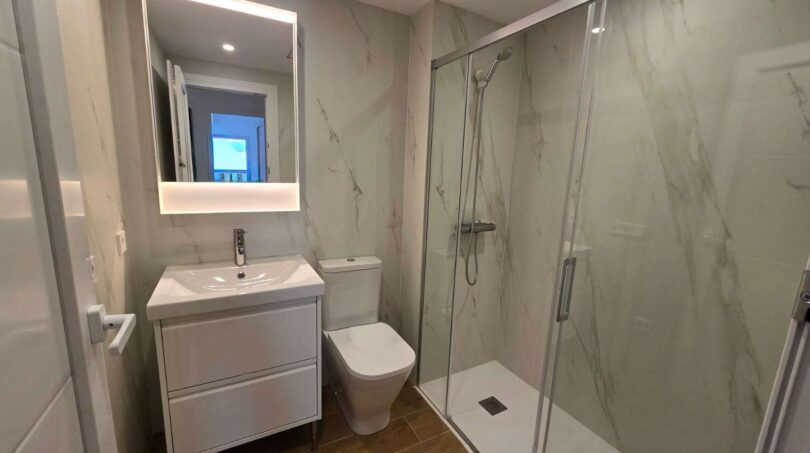 Modernes Badezimmer mit Marmorfliesen, Glasdusche, WC und beleuchtetem Spiegel im Hafenapartment Portocolom
