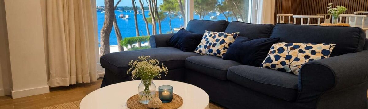 Helles Wohnzimmer mit dunkelblauem Sofa und Panoramafenster mit Meerblick auf den Hafen von Portocolom