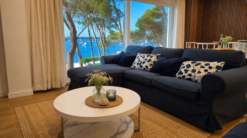 Helles Wohnzimmer mit dunkelblauem Sofa und Panoramafenster mit Meerblick auf den Hafen von Portocolom