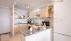 Cocina luminosa con frentes de madera, encimera de granito, isla de cocina y refrigerador blanco en apartamento en primera línea de mar Port Adriano