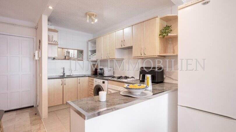 Cocina luminosa con frentes de madera, encimera de granito, isla de cocina y refrigerador blanco en apartamento en primera línea de mar Port Adriano
