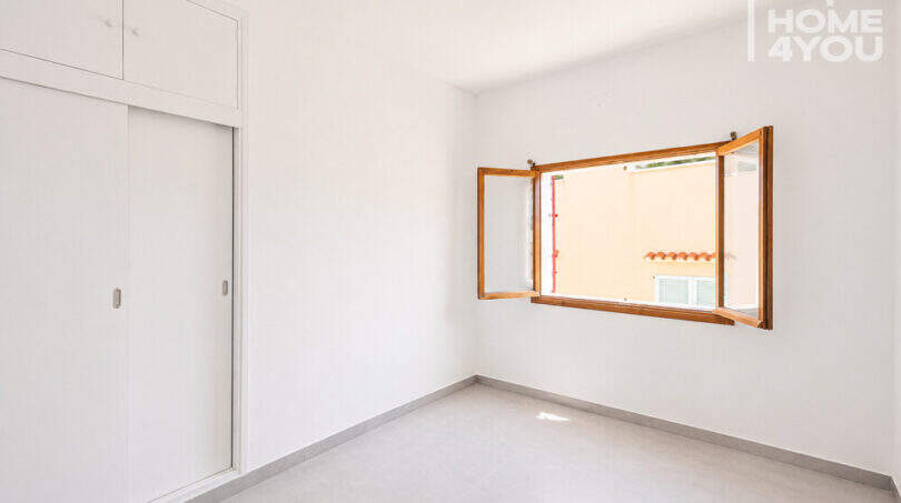 Helles, leeres Schlafzimmer mit Einbauschrank, weißen Wänden, Fliesenboden und geöffnetem Holzfenster zum Innenhof.