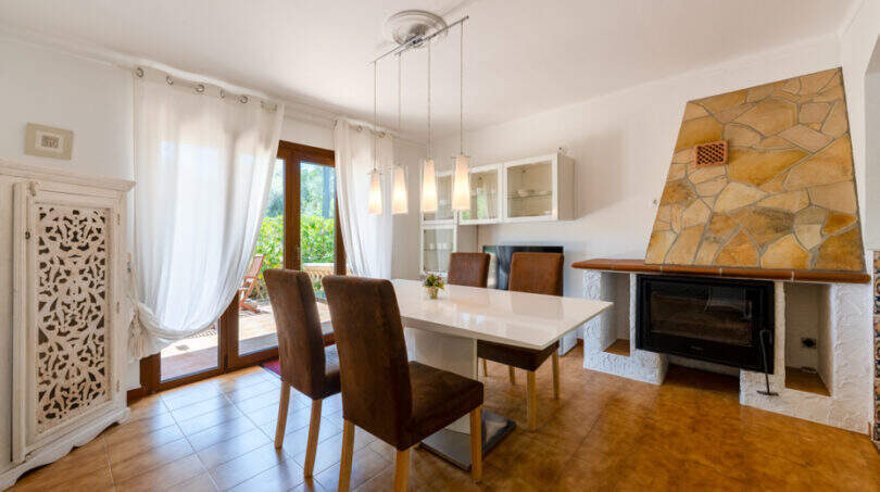 Helles Esszimmer mit weißem Tisch, braunen Stühlen, Naturstein-Kamin und Zugang zur Sonnenterrasse in Cala Murada.