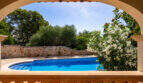 Vista a través de un arco de medio punto hacia una piscina ovalada con borde de mosaico, un muro de piedra natural y una adelfa en flor en Cala Murada