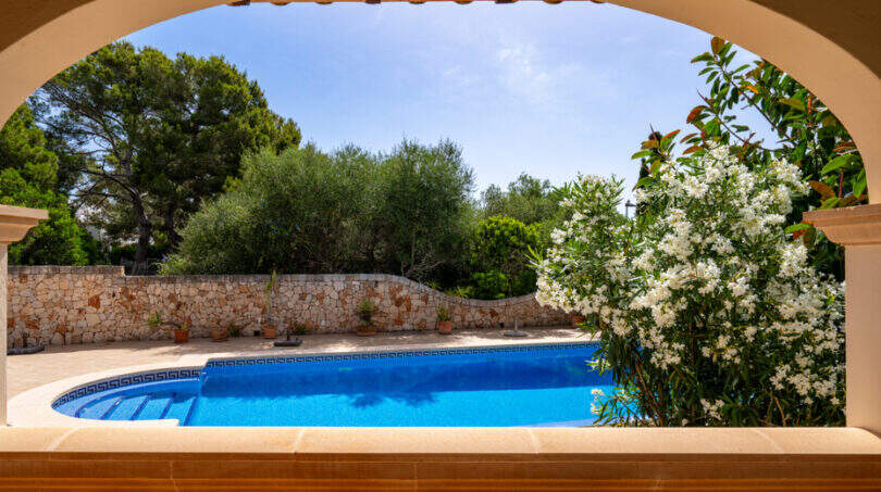 Vista a través de un arco de medio punto hacia una piscina ovalada con borde de mosaico, un muro de piedra natural y una adelfa en flor en Cala Murada