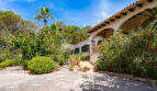 Vista exterior de la Villa Cala Murada: entrada de estilo mediterráneo con arcadas, escalinata y un exuberante jardín con palmeras y plantas con flores.