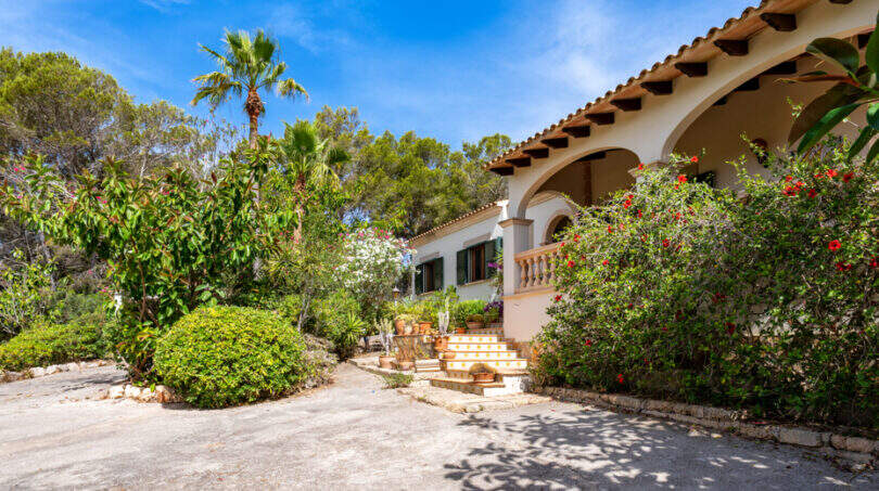 Vista exterior de la Villa Cala Murada: entrada de estilo mediterráneo con arcadas, escalinata y un exuberante jardín con palmeras y plantas con flores.