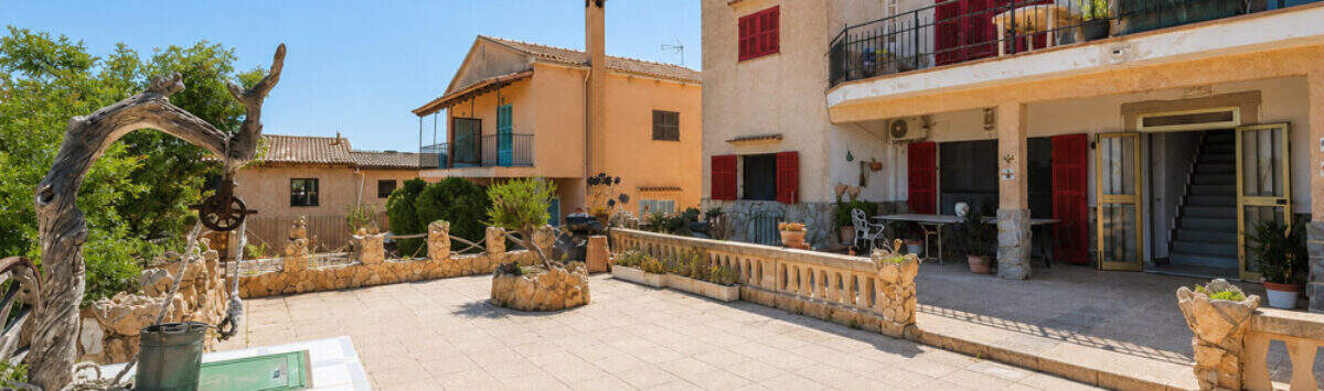 Gepflasterte Terrasse mit Steinbalustrade vor mediterranem Wohnkomplex mit roten Fensterläden in Cala Llombards