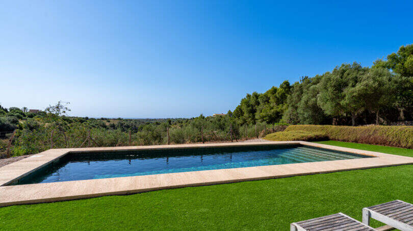 Rechteckiger Außenpool mit Natursteinrandung, gepflegtem Rasen und Meerblick, umgeben von Olivenbäumen in Cales de Mallorca.