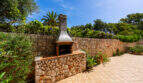 Zona de barbacoa exterior con muros de piedra, chimenea y baldosas de terracota, rodeada de exuberante vegetación, villa familiar Cala Murada