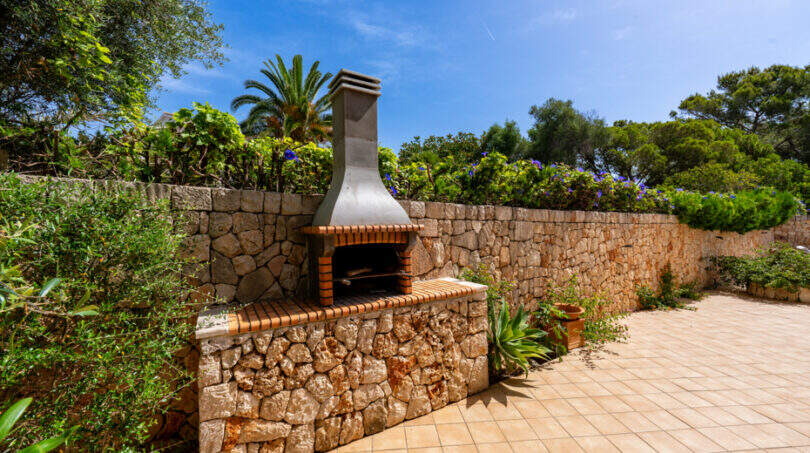 Zona de barbacoa exterior con muros de piedra, chimenea y baldosas de terracota, rodeada de exuberante vegetación, villa familiar Cala Murada
