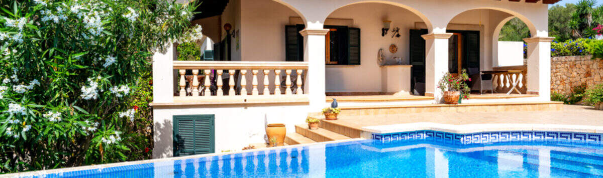 Villa de estilo mediterráneo en Cala Murada con una gran piscina azul, arcadas y un exuberante jardín bajo un cielo azul.