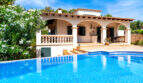 Villa de estilo mediterráneo en Cala Murada con una gran piscina azul, arcadas y un exuberante jardín bajo un cielo azul.