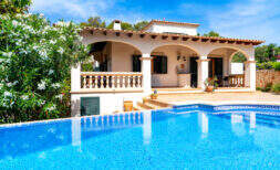 Mediterrane Villa in Cala Murada mit großem blauen Pool, Arkaden und üppigem Garten unter blauem Himmel.
