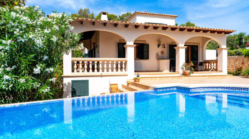 Villa de estilo mediterráneo en Cala Murada con una gran piscina azul, arcadas y un exuberante jardín bajo un cielo azul.