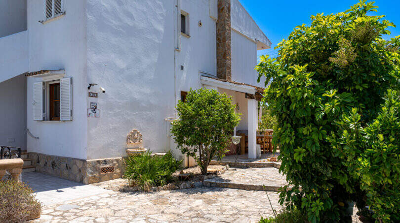 Weißes Erdgeschossapartment in Cala Murada mit Natursteinpflaster, üppigem Grün und überdachter Terrasse.