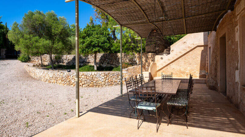 Überdachte Terrasse mit Esstisch aus Schmiedeeisen, Natursteinmauer und Orangenbäumen auf Finca in Mallorca.