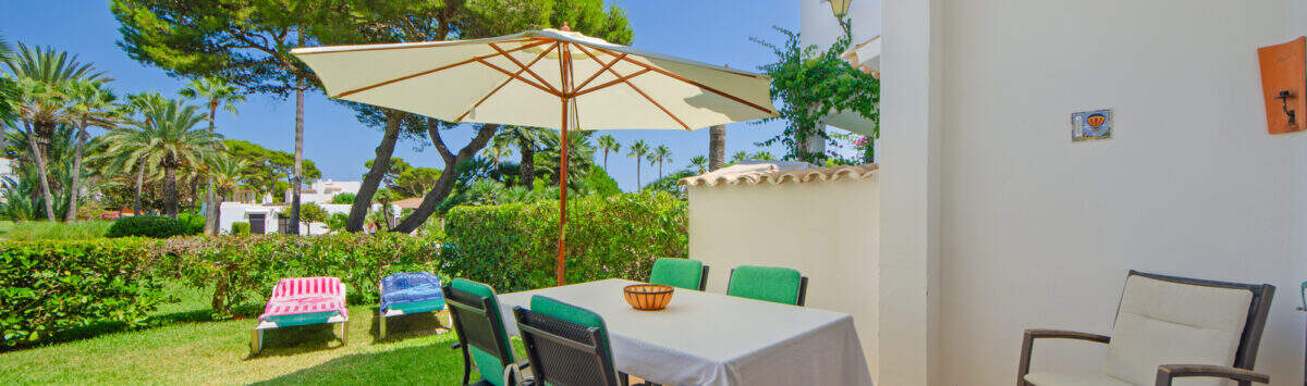 Sonnige Terrasse mit Esstisch und Sonnenschirm, Blick auf gepflegten Garten mit Liegestühlen, Cala d'Or