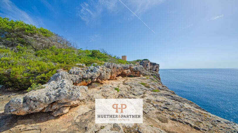 Felsige Klippe in 1. Meereslinie mit blauem Meer, Grünvegetation und historischem Wachturm in Porto Cristo