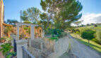 Mediterrane Finca Son Macià: Steinmauer, Pergola mit Arkaden, großer Pinienbaum und gepflegte Gartenanlage mit Zufahrt.