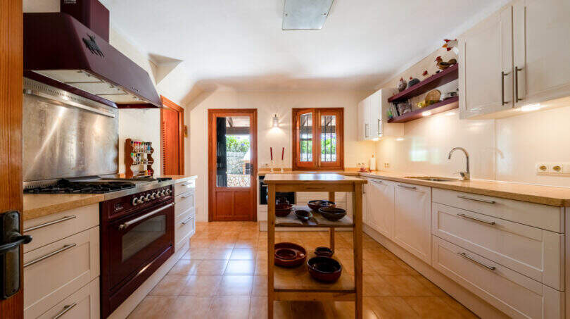 Amplia cocina rústica con cocina de color burdeos, isla de cocina y armarios blancos en Villa Cala Murada