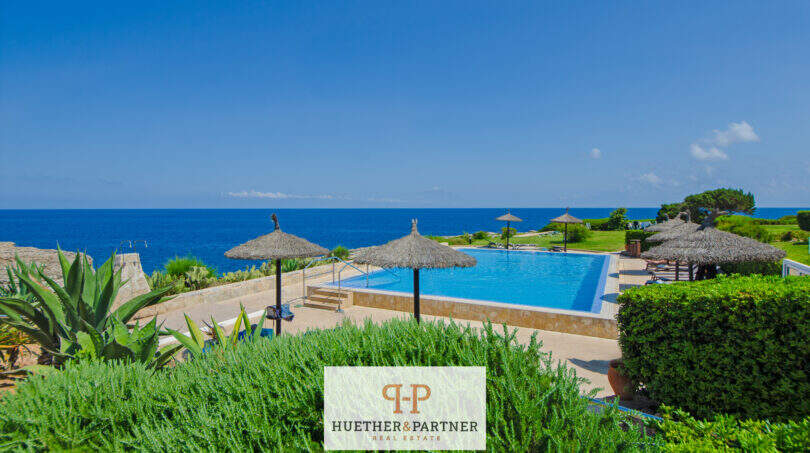 Infinity-Pool mit Meerblick, Strohdach-Sonnenschirme und gepflegte Gartenanlage einer Apartmentanlage in Cala d'Or.