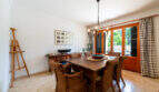 Comedor luminoso de la villa de Cala Murada, con mesa de madera, sillas de mimbre, un caballete y una puerta que da al jardín.
