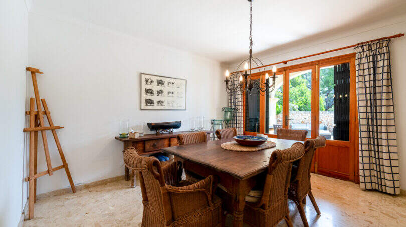 Comedor luminoso de la villa de Cala Murada, con mesa de madera, sillas de mimbre, un caballete y una puerta que da al jardín.